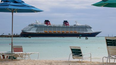 A Carnival cruise ship sails on blue water, seen from a lively beach lined with colorful lounge chairs and umbrellas.