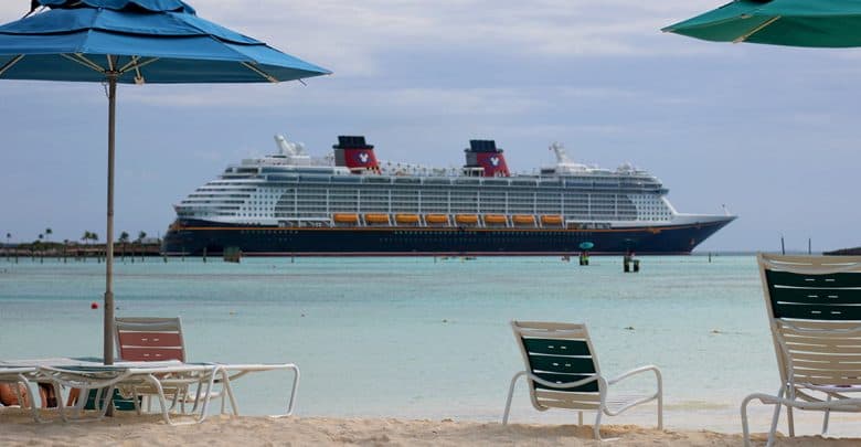 A Carnival cruise ship sails on blue water, seen from a lively beach lined with colorful lounge chairs and umbrellas.