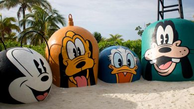 Giant beach buoys featuring Mickey, Pluto, Donald, and Goofy faces brighten a palm-lined shoreline at Disney’s Castaway Cay.