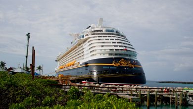 A cruise liner awaits guests at the Disney Cruise Line port, lush landscapes and shimmering blue sea setting the scene.