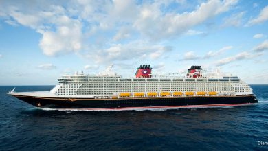 Disney Cruise Line ship with iconic red funnels and yellow lifeboats sailing at sea beneath a partly cloudy sky.