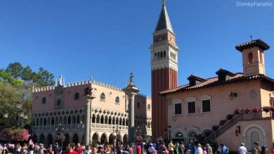 A crowd gathers in front of Italian-style buildings and a tall bell tower, with clear blue skies above. Pink flowers line a staircase on the right. Trees are visible in the background.