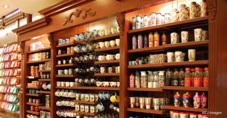 Brightly lit shelves stocked with Disney-themed mugs, colorful souvenir cups, bottles, and snacks at a theme park gift shop.