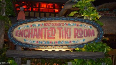 A wooden sign surrounded by tropical plants reads "Walt Disney’s Enchanted Tiki Room" in colorful, stylized letters, illuminated at night at Walt Disney World with a building and warm lighting in the background.