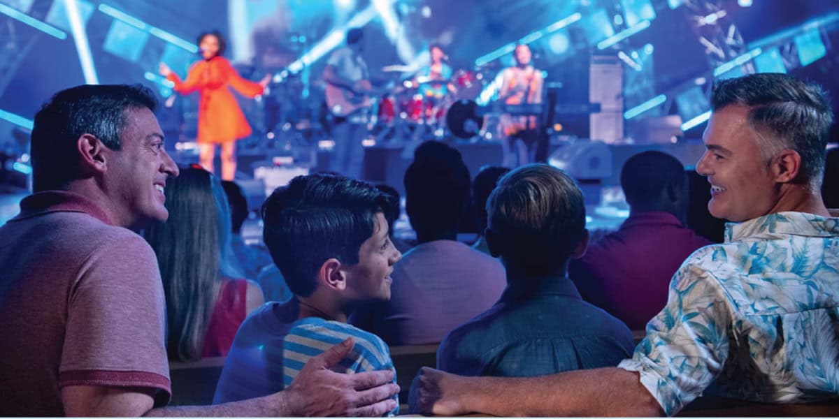 Two men and two boys sit together in an audience, smiling and enjoying a live music performance on a brightly lit stage—an experience as delightful as planning your visit with the Walt Disney World crowd calendar.