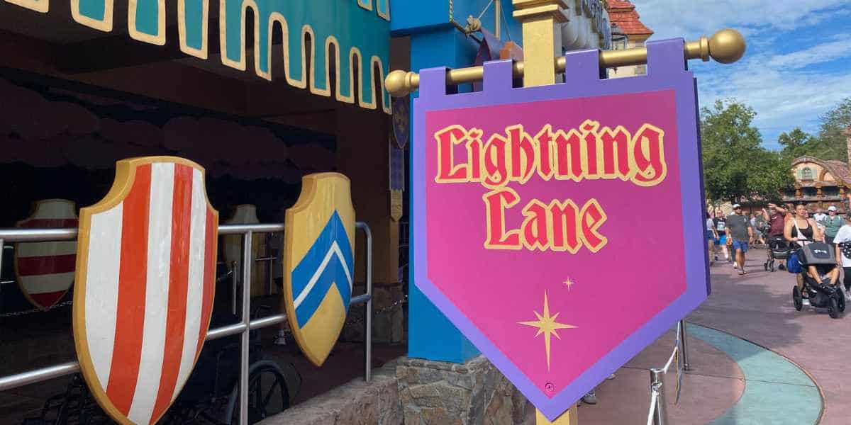 A purple sign reading "Lightning Lane" stands near a ride entrance adorned with medieval-style shields and decorations, indicating the access point for guests using Lightning Lane Passes; people are walking and pushing strollers in the background.