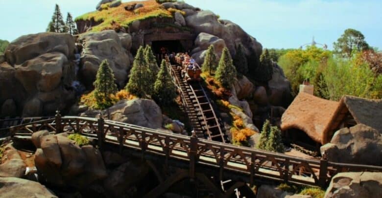 A wooden roller coaster track, reminiscent of Magic Kingdom rides, winds through a rocky, forested hill. A train full of riders descends out of a tunnel amidst trees and shrubs, while a wooden bridge crosses over the lower track. The sky remains clear and blue.