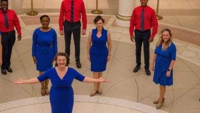 A group of eight, four women in blue dresses and four men in red shirts with black pants, stand in a semi-circle on a patterned floor, beaming as if capturing the joy of touring EPCOT. They appear to be performing or posing for a photo indoors.