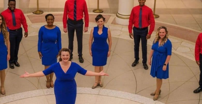 A group of eight, four women in blue dresses and four men in red shirts with black pants, stand in a semi-circle on a patterned floor, beaming as if capturing the joy of touring EPCOT. They appear to be performing or posing for a photo indoors.