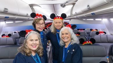 Four smiling women wearing Mickey Mouse ear headbands stand inside an airplane cabin. Despite recent flight cancellations, the mood is cheerful and festive, with Mickey Mouse ears on every headrest adding to the fun atmosphere.