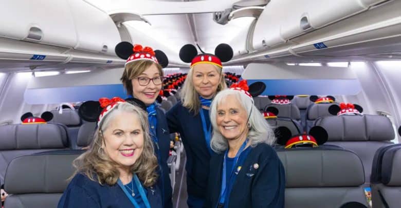 Four smiling women wearing Mickey Mouse ear headbands stand inside an airplane cabin. Despite recent flight cancellations, the mood is cheerful and festive, with Mickey Mouse ears on every headrest adding to the fun atmosphere.