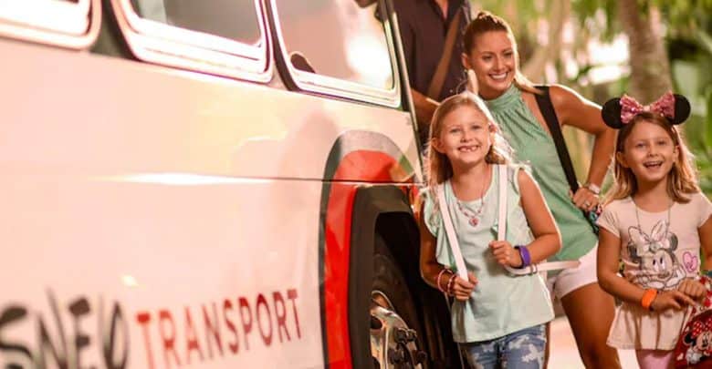 A smiling adult and two young girls, one wearing Mickey Mouse ears, stand next to a Disney Transport bus with red and white colors on a sunny day, enjoying magical moments together despite recent flight cancellations.
