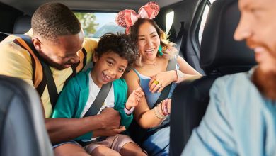 A smiling family sits together in a car, undeterred by flight cancellations; a man buckles in a laughing child while a woman in sparkly pink mouse ears sits beside them, all appearing excited and joyful.