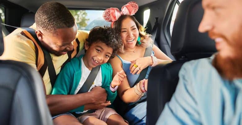 A smiling family sits together in a car, undeterred by flight cancellations; a man buckles in a laughing child while a woman in sparkly pink mouse ears sits beside them, all appearing excited and joyful.