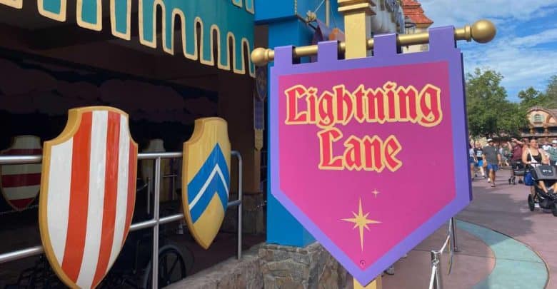 A purple "Lightning Lane" sign with gold and pink lettering marks a Disney Genie+ entrance, set near medieval-style shields at a theme park. In the background, people walk and push strollers on a sunny day.