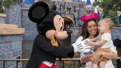 A woman holding a smiling baby stands next to Mickey Mouse, who is reaching out playfully toward the child in front of a castle at Disneyland—highlighting the joy and excitement of keeping kids safe at Disney World.