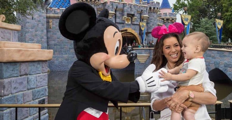 A woman holding a smiling baby stands next to Mickey Mouse, who is reaching out playfully toward the child in front of a castle at Disneyland—highlighting the joy and excitement of keeping kids safe at Disney World.