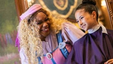 A smiling woman dressed as a fairy godmother in a pink and blue costume chats with a young girl in a salon cape, both appearing happy inside a decorated room, sharing excitement about Walt Disney World trip planning.