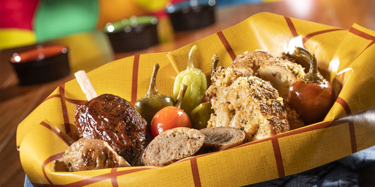 A basket lined with yellow paper holds assorted grilled foods—a meatball on a stick, sausage slices, stuffed peppers, and a whole cauliflower—perfect for fueling up before diving into Disney World planning tips. Colorful condiments blur in the background.