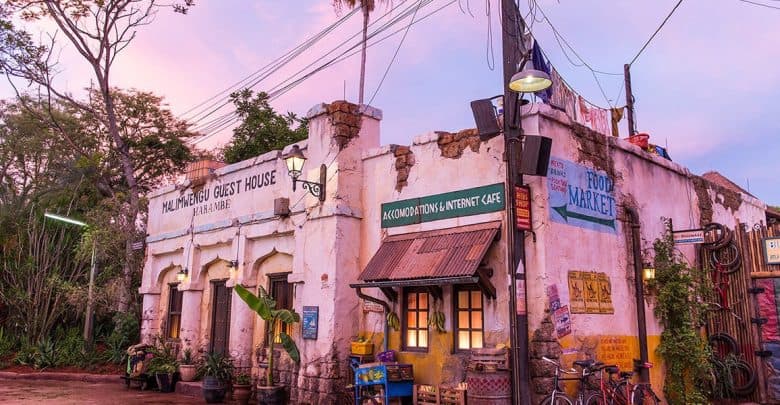 A rustic, weathered building with faded signs reading "Guest House" and "Food Market" sits beneath a pink-purple sky, surrounded by greenery, bicycles, and vintage decor—evoking old-world charm perfect for those seeking to stay cool at Disney World.