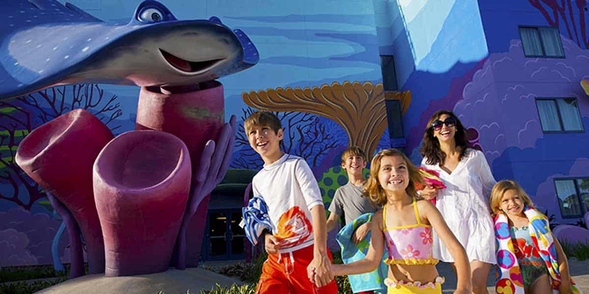 A smiling family in swimsuits walks outside a brightly painted building with large, colorful ocean-themed decorations, including a big friendly blue stingray and coral sculptures—an image straight out of glowing Disney Resort Reviews.