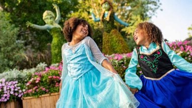 Two young girls dressed as princesses joyfully hold hands and smile in a garden filled with colorful flowers, beneath topiary figures of Frozen's Elsa and Anna—a magical scene often found during the busiest times at Walt Disney World.