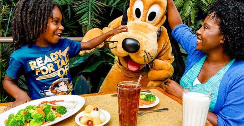 A child and adult smile and interact with a person in a Pluto costume at a restaurant table—showing that knowing what to avoid at Disney World means you can relax, enjoy delicious food, and soak in the lush greenery around you.