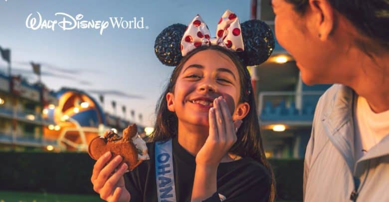 A young girl wearing Mickey Mouse ears and a sash smiles while holding a Mickey-shaped ice cream sandwich, sitting next to an adult at Walt Disney World. They’re clearly enjoying the magic—a reminder to focus on fun and know what to avoid at Disney World.