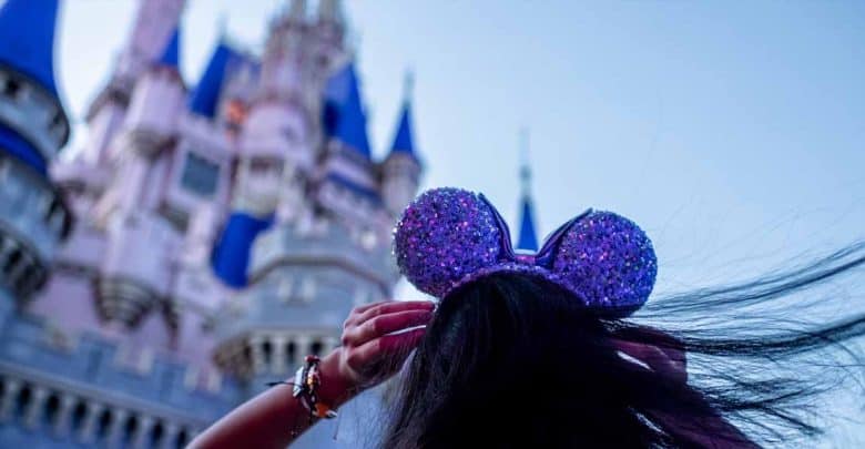 A person wearing sparkly Mickey Mouse ears faces a large, pink and blue fairytale castle, their long hair blowing in the wind. The bright, whimsical scene hints at theme park adventures and Magic Kingdom tips for making memories.