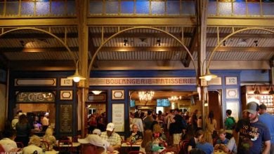 A bustling indoor café named "Boulangerie Patisserie," nestled among the quick service locations inside the Disney theme parks, with people sitting at tables and standing in line at the counter. The décor boasts warm lighting and an ornate ceiling, crafting a lively and inviting atmosphere.