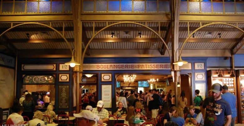 A bustling indoor café named "Boulangerie Patisserie," nestled among the quick service locations inside the Disney theme parks, with people sitting at tables and standing in line at the counter. The décor boasts warm lighting and an ornate ceiling, crafting a lively and inviting atmosphere.