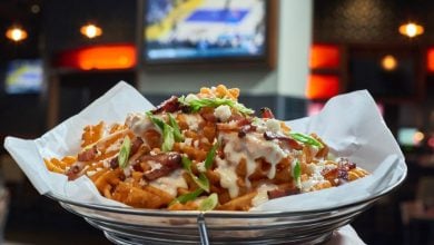 A basket of waffle fries topped with cheese sauce, bacon bits, and chopped green onions sits on white paper lining. In the background, a blurred TV screen displays a basketball game in a dimly lit setting—just the kind of scene you'd find at the best sports bars at Disney World.