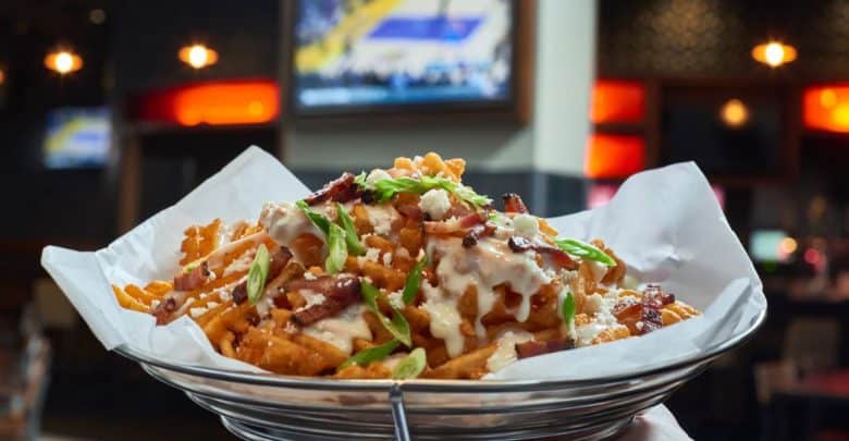 A basket of waffle fries topped with cheese sauce, bacon bits, and chopped green onions sits on white paper lining. In the background, a blurred TV screen displays a basketball game in a dimly lit setting—just the kind of scene you'd find at the best sports bars at Disney World.