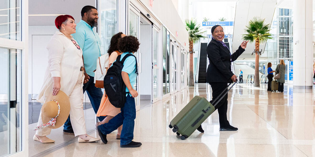 A smiling airport staff member leads a family with luggage through a bright, modern terminal with palm trees inside, helping them avoid holiday travel disasters. The family of four looks cheerful and ready to travel.