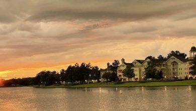 A serene sunset over a lakeside neighborhood with a large, elegant building and trees lining the shore. The sky is filled with dramatic clouds, and the sun is just above the horizon, casting a warm glow over the water—a perfect scene reminiscent of running at Disney’s picturesque settings.