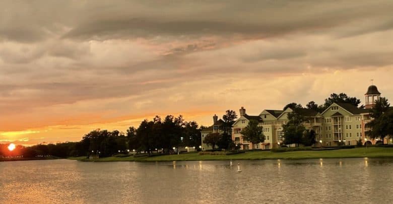 A serene sunset over a lakeside neighborhood with a large, elegant building and trees lining the shore. The sky is filled with dramatic clouds, and the sun is just above the horizon, casting a warm glow over the water—a perfect scene reminiscent of running at Disney’s picturesque settings.