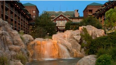 A scenic view of a rustic lodge-style building with a green roof, surrounded by trees. In the foreground, a gentle waterfall cascades over rocks into a small pond, creating a tranquil natural setting—perfect for relaxing at Disney's enchanting escape.