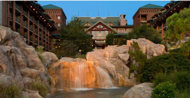 A scenic view of a rustic lodge-style building with a green roof, surrounded by trees. In the foreground, a gentle waterfall cascades over rocks into a small pond, creating a tranquil natural setting—perfect for relaxing at Disney's enchanting escape.