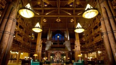 The grand hotel lobby at Disney's Wilderness Lodge boasts tall wooden columns and large, glowing chandeliers hanging from the high ceiling. The open, spacious area features seating arrangements and rustic timber architecture, creating a warm, inviting atmosphere reminiscent of an enchanting retreat.