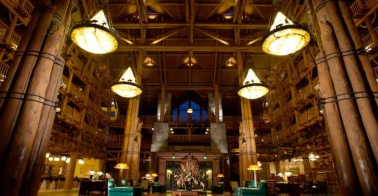 The grand hotel lobby at Disney's Wilderness Lodge boasts tall wooden columns and large, glowing chandeliers hanging from the high ceiling. The open, spacious area features seating arrangements and rustic timber architecture, creating a warm, inviting atmosphere reminiscent of an enchanting retreat.