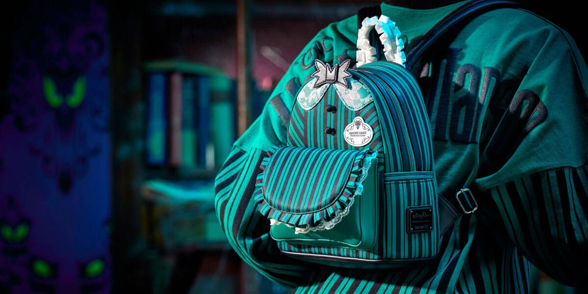 A person wearing a teal and navy striped sweater carries a matching mini backpack with ruffles, butterfly decorations, and a metal Haunted Mansion tag—perfect for those seeking Disney World planning tips—in a dimly lit room with blurred shelves behind them.
