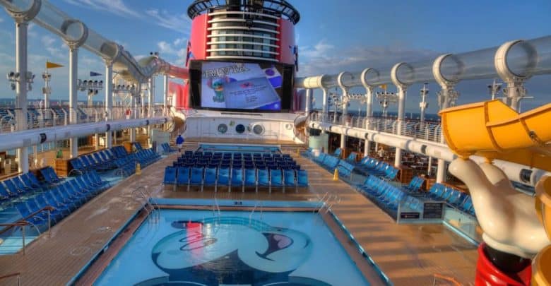 A cruise ship's deck with a pool in the foreground featuring a large image of Mickey Mouse. Numerous blue lounge chairs surround the pool. A large screen shows a movie, and a yellow water slide curves into view on the right. The sky is partly cloudy, adding charm to this Disney Cruise Line experience.