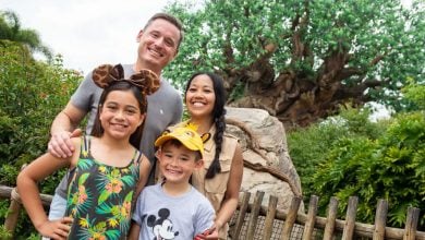 A smiling family of four poses in front of a lush, leafy tree at a theme park. The children wear Mickey Mouse-themed hats and shirts, while the parents stand behind them, all looking happy—ready for more Disney World planning tips.