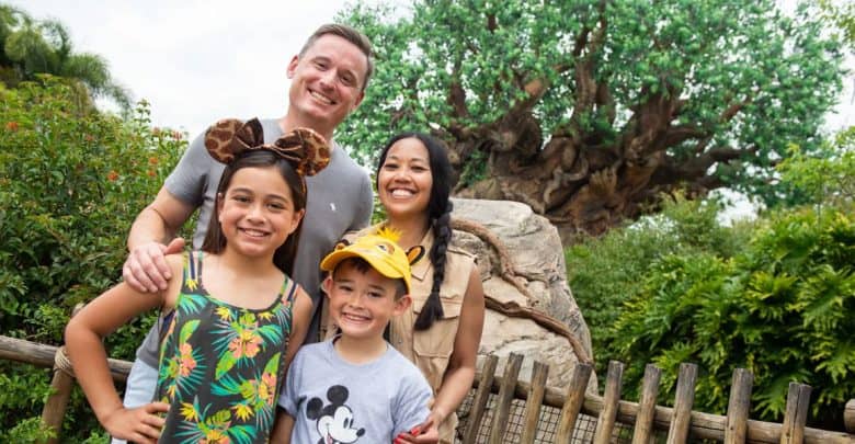 A smiling family of four poses in front of a lush, leafy tree at a theme park. The children wear Mickey Mouse-themed hats and shirts, while the parents stand behind them, all looking happy—ready for more Disney World planning tips.
