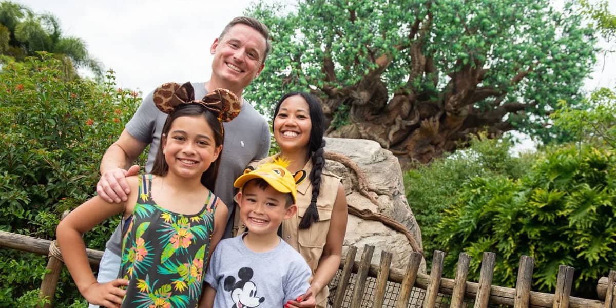 A smiling family of four poses in front of a lush, leafy tree at a theme park. The children wear Mickey Mouse-themed hats and shirts, while the parents stand behind them, all looking happy—ready for more Disney World planning tips.