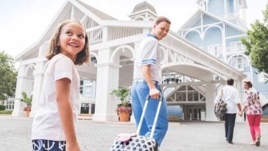 A smiling young girl and a man pull a polka-dot suitcase outside a large white and blue resort building, ready to start their adventure with handy Disney Planning Tips, while two adults walk ahead toward the entrance.