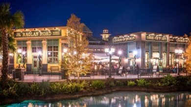 At Disney Springs, the night view of Raglan Road, an inviting Irish pub and Disney restaurant, features a charming green exterior adorned with bright lights. Surrounded by trees and reflected in nearby waters, diners enjoy the ambiance on the patio under twinkling string lights.