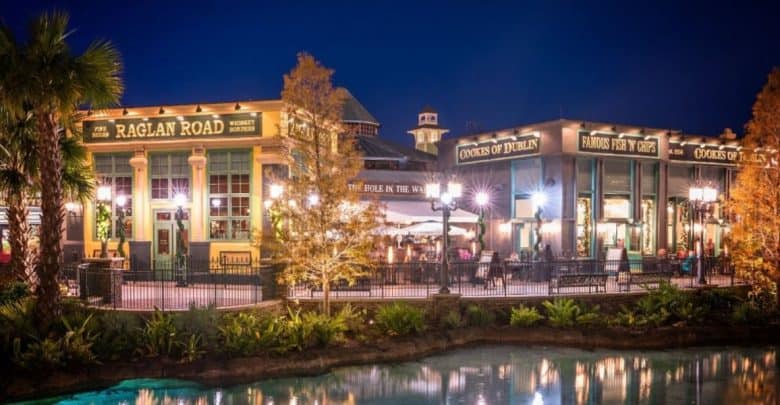 At Disney Springs, the night view of Raglan Road, an inviting Irish pub and Disney restaurant, features a charming green exterior adorned with bright lights. Surrounded by trees and reflected in nearby waters, diners enjoy the ambiance on the patio under twinkling string lights.
