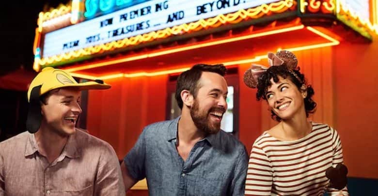 Three people sit in front of a brightly lit theater marquee at Walt Disney World.