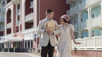 A man and woman, dressed in vintage 1920s attire, walk arm in arm outside colorful, resort-style buildings at one of the Best Disney Resorts for Evening Activities on a sunny day.